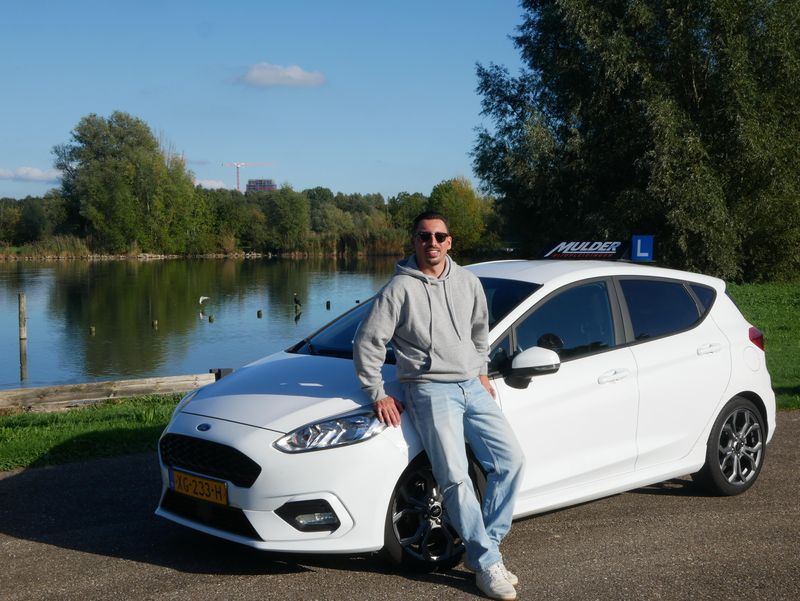 Rijinstructeur Chris Mulder leunt tegen de witte Ford Fiesta lesauto bij een meer in IJsselstein
